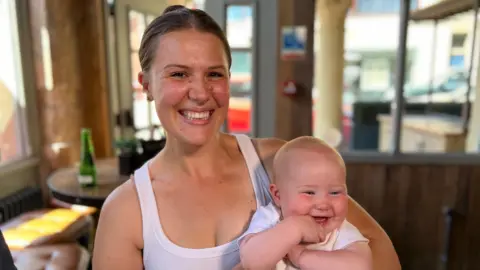 Hollie Benneyworth is pictured smiling widely while holding a giggling baby. Both are wearing white tops and Hollie has her brown hair tied back in a bun with a centre parting. Mother and baby appear to be sitting in a cafe.