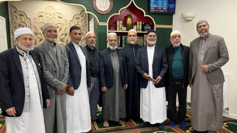 Jon Wright/BBC Nine men standing in a line at the front of a mosque. They are wearing traditional Muslim robes (called Thobes)