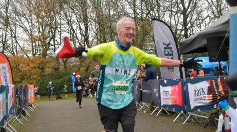 Mike Barnes Mike Barnes has his arms stretched out as his crosses the finish line during a rave. He is wearing a yellow and green running jersey with his name on. His daughter's name is on the back. 