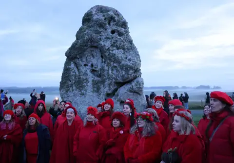 EPA Women dressed all in red stand in front of a Neolithic stone singing.