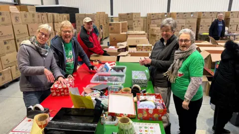 Madeleine McClintock Five volunteers smiling at the camera. They are standing around a table which has wrapped shoeboxes on. 