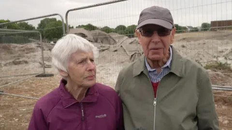 Jennifer and Ray Hessan stood together in front of the building site. The area has a metal fence around it. 