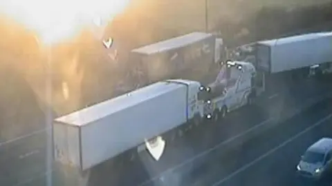 Traffic England Three lorries, two white and one a darker colour on the road. One is being attached to a recovery vehicle.