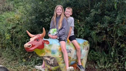 A young boy and girl are sat on a multi-coloured statue of a cow, in front of a green, leafy background. The girl is sat at the front and the boy at the back of her. The cow has a red face and horns, with blue and green print on its body. 