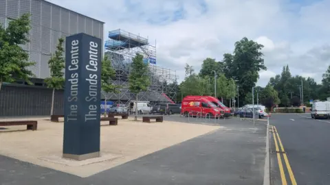 A general view of the entrance to The Sands Centre in Carlisle. A black pillar has the name of the venue written on each side in silver. The main building is behind the pillar and on the right there is an area of the building surrounded by scaffolding.