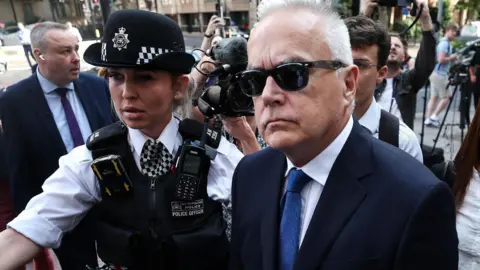 Police officer next to Huw Edwards outside court. He is wearing a dark suit, light blue tie and dark sunglasses