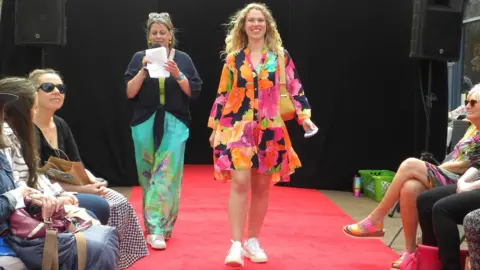 Discover Daventry Woman with long blond hair wearing a multi-coloured dress in light fabric and an orange handbag. She also has white plimsolls. She is walking down a red carpet. There is a woman behind her wearing a black T-shirt and blue trousers with a floral design above white plimsolls, who is reading from a piece of white paper into a microphone. There are people sitting on either side of the carpet.