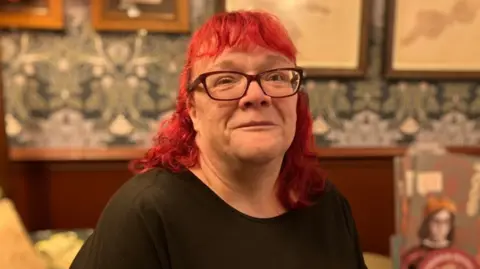 A woman with shoulder length red hair and dark glasses. She is smiling at the camera