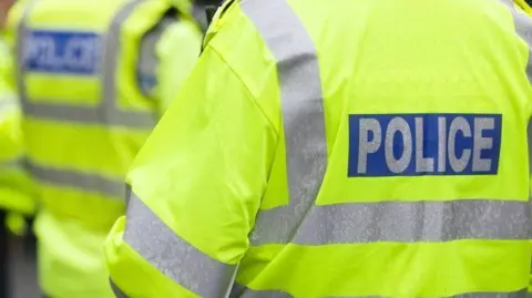 Three police officers standing outside with their backs to the camera. They are all wearing yellow fluorescent jackets which have silver stripes on the back and arms and writing, which says POLICE on the back. 