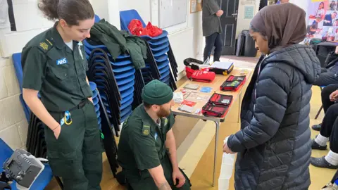 Two paramedics wearing forest green uniform, decorated with an NHS badge, the SCAS logo and shoulder panels are talking to young people at the event. The female paramedic with brown hair tied in a ponytail has her hands in her pockets and looks towards the floor, while her male colleague, who wears a green beanue hat and has a ginger beard, is kneeling on the floor while he talks. Standing in front of them both is a young woman, who wears a brown hijab and long black hooded puffer coat, she is looking towards the floor where the male paramedic is.