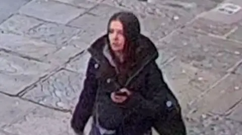 A white girl, about 5ft 4ins tall, with brown hair, wearing a grey hoodie, dark jogging bottoms, black Nike Air Force trainers, and carrying a black bag.