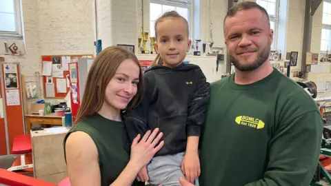 Yana and her husband Taras, who wear green club shirts, hold up their son Leo in the gym 