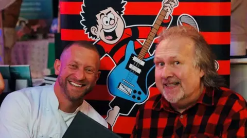 La Vida Liverpool & Jade Andreas Dave Critchley, wearing chef whites, sits next to Nigel Parkinson, who is wearing a red and black plaid shirt. A banner with a picture of Dennis the Menace, a cartoon boy, playing an electric guitar is behind them. 