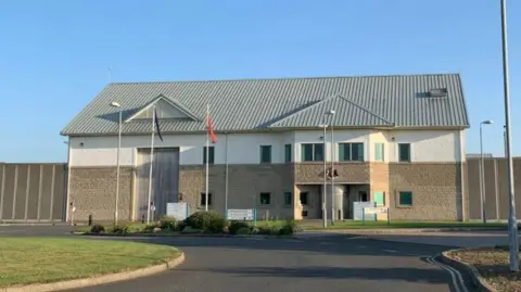 The exterior of the prison, a large brown building, with CCTV, all around.