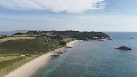 A birds-eye view of a beach on Herm. There is a green cliff area with coastal path running through it and white sand along the beach area. On the right is clear blue sea with a rock at the centre. 