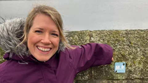 A woman with blond hair is smiling and wearing a purple coat. She is standing by a wall with a small sign which says the letters M and P