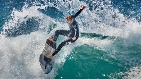 Peter Hicks A man is surfing on a blue wave with an arc of white water behind him. It's Finlay surfing on a Cornish beach. 