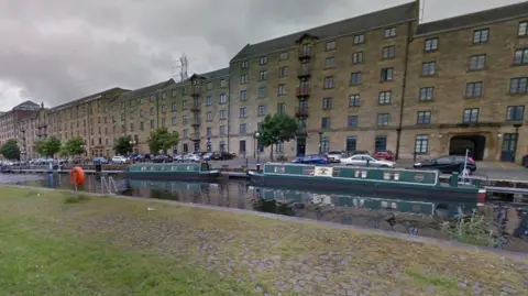 A canal with several small barges and boats moored there. There are flats and cars alongside it