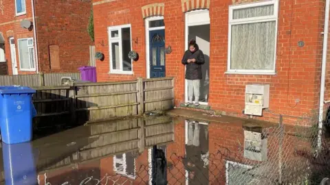 A flooded garden with a woman standing in a doorway cut off by water taking pictures of the garden.