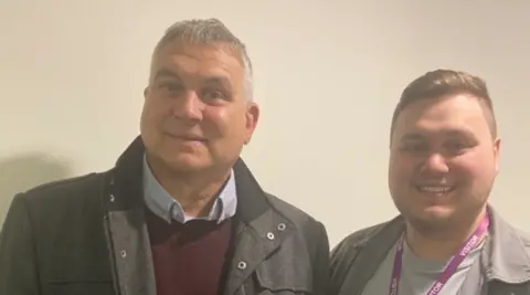 A man with grey hair wearing a dark top and purple jumper is on the left in front of a wall. A younger man with brown hair and wearing a lanyard is on the right.