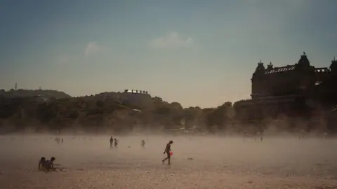 Emma Warley Picture of Warley's - which shows many people walking and digging on Scarborough's South Bay beach. A mist comes up over the beach whilst trees and Scarborough's buildings can be seen looking down onto the beach.