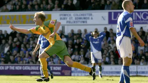 Getty Images Chadwick, celebrating scoring in the Yellow and Green of Norwich City.  He has one finger raised in celebration, as an Ipswich Town player, in full blue and white kit, looks dejected at conceding at Portman Road. 