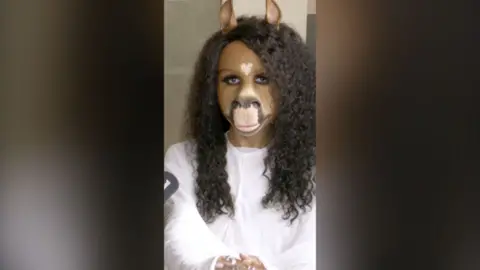 horsegiirL wearing her signature mask and staring directly at the camera. She's wearing a white top.