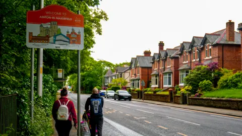 PA Media Two people walk past a sign for Ashton-in-Makerfield on an overcast day.