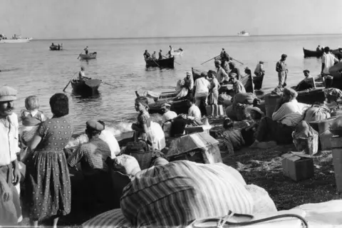 AP a black and white photo showing households with gathered bags in a beach. Some of them are rowing in distances in small woody wire bars