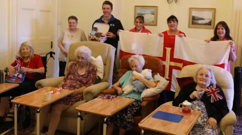 Four people sat in chairs smiling at the camera. There is five people stood behind them, three holding a flag and one holding a hamper.