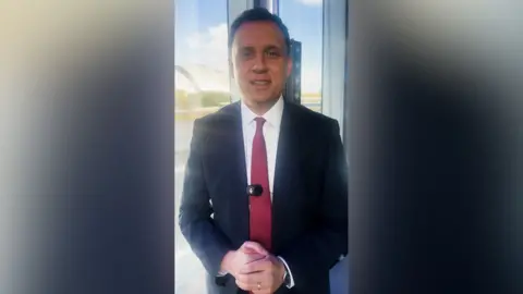 Anas Sarwar smiles at the camera as he stands in front of a wide window. He has black hair and brown eyes and is wearing a red tie, white shirt and black suit.
