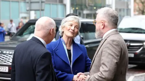 Pacemaker Catherine Connolly, a woman with grey hair, wearing blue jacket and black top, shakes hands with a man with brown jacket, grey hair and beard and brown jacket. To the left is a man with short grey hair and black jacket.