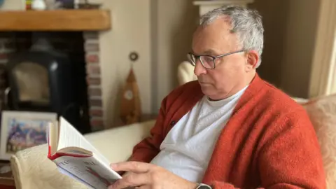 Andrew in a white T-shirt and orange cardigan sitting in an armchair reading at home. 