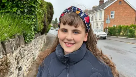 A girl is standing on a pavement next to a stone wall and green bushes. She is wearing a dark blue quilted jacket and a colorful headband with red, yellow, and white flowers. In the background, there is a residential street with houses, parked cars.