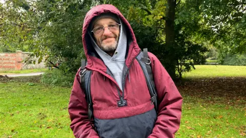 Michael Stevenson wearing a waterproof coat with the hood up, glasses, and carrying a rucksack, standing in a green area with a tree behind him