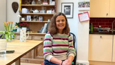 Toby's mother, Claire, is sitting next to a table with a vase of flowers and a jug with spots on. She has shoulder-length brown hair and is wearing a striped green, purple and white jumper. There is a bookcase behind her and a kitchen in the background.