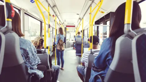 Getty Images Passengers sitting on a bus seen from behind, with one woman standing