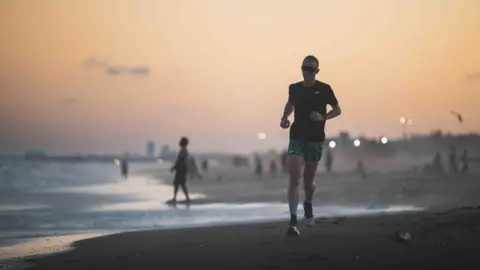 James Poole Mr Robinson running along the beach at sunset. He is wearing green and black patterned shorts and a black t-shirt. Behind him are people enjoying the beach on the shoreline. The sky is dim but still glowing orange.