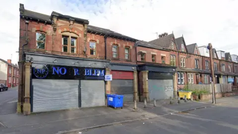 A Street View image of the shop, which has its grey shutters down. The store name is written in blue letters but is missing the P and O. A blue bin has been left to the right of the store.