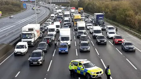 A police car blocks the flow of traffic across six lanes of the M25 