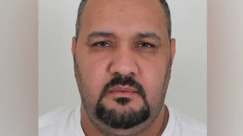 A man with a black beard and partly grey hair is looking at the camera. He is wearing a white T-shirt in front of a grey background.