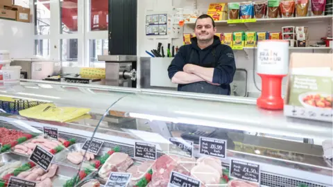 Kenny Brown Butcher Scott Donnelley with his arms folded, smiles behind his meat counter in front of mince, sausages and bacon 