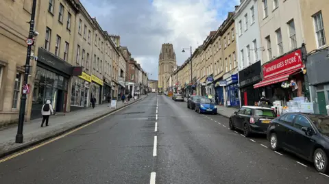 A photograph of Park Street, with the Wills Building at the top of the road in the centre. The sky is cloudy and the road is empty apart from a few parked cars. There are shops lining the street including a homewares store and a vintage clothes store.