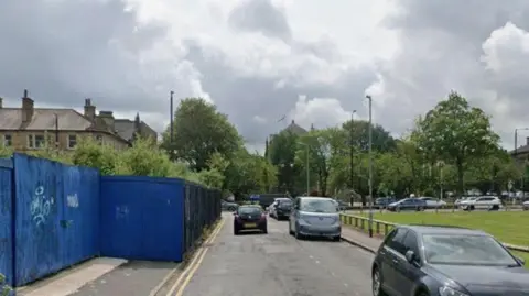 A road filled with parked cars, with one car driving away from camera. Green space is on the right, with a boarded up area on the right. 