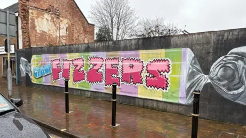 A long wall beside a wet sidewalk shows a mural painted like a wrapped candy. It has pastel stripes and large pink letters spelling “FIZZERS.” 