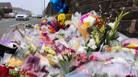 A large roadside memorial made up of many bouquets of flowers arranged along a pavement beside a stone wall.
