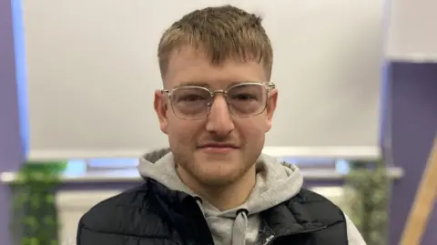 Will Postgate is wearing large silver rimmed glasses, a grey hoodie with a black gilet over the top. He has light brown stubble and light brown hair.