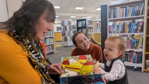 A baby and two women look at a picture book together in a library. The two women are looking at the baby. There are rows of books on bookshelves behind the trio. 