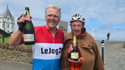 Eddie McGourley and Stephen Foster stand in front of a sign reading 'John O'Groats' with a bottle of champagne each. The sky is cloudy and blue and there is a white-and-grey building behind them. Stephen on the left has grey hair and is wearing a red, white and blue cycling top. Eddie on the right, wears a white cycling helmet and brown jacket buttoned over a red cycling top.