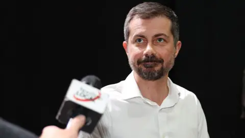 Pete Buttigieg wears a white button down shirt and has a scruffy trimmed beard.